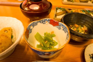 Hafif baharatlı yeşil sebzeler ahşap bir masada çeşitli Japon kahvaltı tabakları ile çevrili renkli bir porselen kasede servis edilir. Sıcak aydınlatma davetkar dokuları ve canlı sunumu geliştirir.