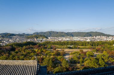 Himeji Kalesi 'nden yükselen manzara sonbahar yeşilliklerinin canlı bir karışımını gösteriyor, geleneksel kiremitli çatılar, ve açık mavi gökyüzünün altında yuvarlanan yeşil tepelere karşı kurulmuş Himeji şehri..