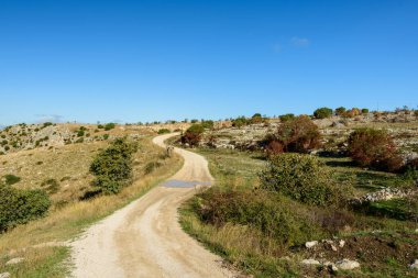 Kıvrımlı toprak yol, dağılmış çalılar, kayalık zemin ve canlı mavi gökyüzü ile güneşli bir platodan geçiyor. Bu da İtalya 'nın Matera kenti yakınlarında huzurlu bir kırsal atmosferi çağrıştırıyor..