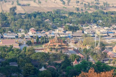 Kamboçya 'nın küçük bir kasabası, yemyeşil ağaçları ve Phnom Sampov yakınlarındaki kuru tarım alanlarıyla çevrili geleneksel bir Budist tapınağı manzarası. Kompozisyon, dini mimarinin, köy hayatının ve
