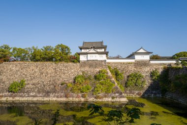 Tarihi taş duvarlar ve Osaka 'daki sakin bir hendeğin üzerinde yükselen beyaz bir Japon kalesi, yemyeşil ve parlak öğleden sonra ışığında yıkanan. Açık mavi gökyüzü huzurlu ve zamansız atmosferi güçlendirir..