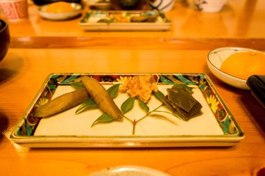 Burdock kökü ve deniz yosunu da dahil olmak üzere çeşitli turşulu sebzeler, renkli bir çiçek sınırının yer aldığı dikdörtgen seramik tepside yenilebilir yapraklarla ustaca düzenlenir. Sıcak ortam aydınlatması bir şeyin üzerindeki dokuları ve zarif sunumu vurgular