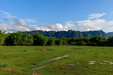 Geniş çimenlik alan, su parçaları ve yoğun yeşil çalılar, engebeli kireçtaşı dağlarına karşı kurulmuş parlak mavi gökyüzü altında. Sahne, Laos kırsalının doğal dokularını ve açık atmosferini yakalıyor..