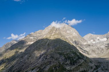 Kayalık alp zirvesi, engebeli, gölgeli bir tepenin üzerinde yükselir. Kar parçaları ve küçük bulutlarla kaplı koyu mavi bir gökyüzü dramatik bir dağ manzarası yaratır..