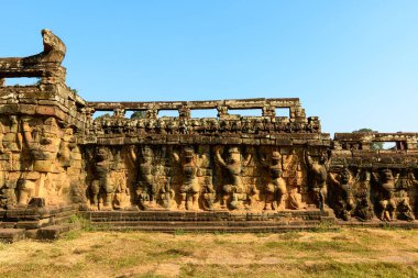 Ilık güneş ışığı, Kamboçya 'nın Angkor kentindeki Filler Terası boyunca mitolojik figürlerin detaylı taş oymalarını aydınlatır. Yıpranmış kumtaşı kabartmaları açık mavi gökyüzüne karşı duruyor, tarihi eserleri vurguluyor.