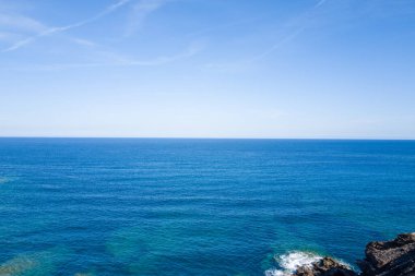 Arjantin yakınlarındaki açık, güneşli bir gökyüzü altında, yumuşak dalgalar ve kayalık bir kıyı şeridiyle ufka uzanan sakin mavi Akdeniz manzarası.