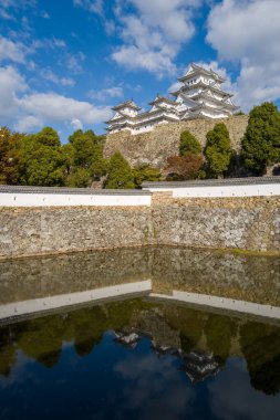 Himeji Kalesi 'nin çarpıcı beyaz kuleleri antik taş duvarların ve yemyeşil yeşilliklerin üzerinde yükseliyor. Çevreleyen hendeğin sakin yüzeyine mükemmel bir şekilde yansıyor. Parlak gün ışığı ve dağınık bulutlar canlı ve sakin bir manzara yaratır..
