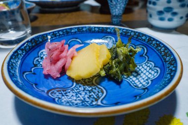 Japon turşulu üç çeşit sebze turşusu. Pembe, sarı ve yeşil. Arkaplanda zarif sofra takımı bulunan, karmaşık bir şekilde düzenlenmiş mavi seramik tabakta. Yakın çekim, canlı renkleri ve zıt dokuları vurgular.