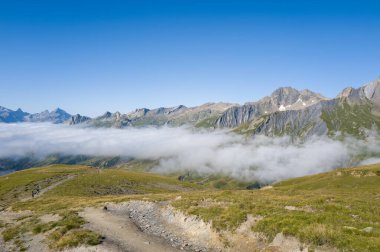Beyaz bulutlar, engebeli dağ zirveleri ve arka planda açık mavi gökyüzü olan yemyeşil bir vadinin üzerinde süzülür. Ön planda, açık ve canlandırıcı bir atmosfer yaratan, dolambaçlı toprak bir patika ve çimenli yamaçlar yer alıyor..