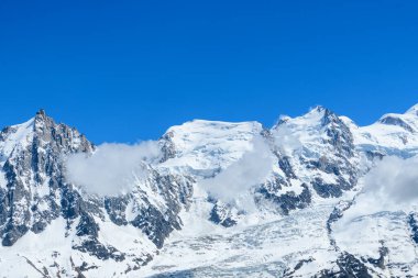 Mont Blanc du Tacul ve Mont Maudit 'in karla kaplı zirveleri, engebeli dağlık arazi boyunca sürüklenen bulut zerreleriyle canlı mavi bir gökyüzü altında keskin bir şekilde yükselir..