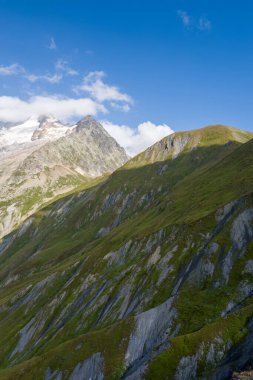 Keskin çimenli tepeler ve kayalık yamaçlar Val dAoste dağlarındaki berrak mavi gökyüzünün altında uzak buzullarla kaplı bir zirveye doğru yükseliyor..