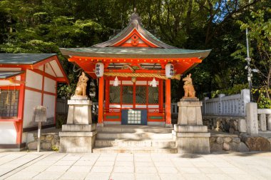 Japonya 'nın Kobe kentindeki geleneksel bir Shinto türbesi pavyonunda kalın kırmızı kirişler, yeşil kafes kapılar ve iki taş koruyucu aslan köpek heykeli bulunuyor. Güneş ışığı mimariyi aydınlatır ve taş basamaklarda ve avluda canlı gölgeler bırakır..