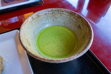 Canlı yeşil kibrit çayıyla dolu el yapımı bir seramik kâse siyah bir tepside duruyor, köpüklü yüzeyi parlak kırmızı bir masa üzerinde parlıyor..