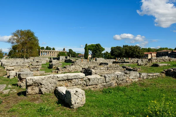 Yıpranmış taş, İtalya 'nın Paestum kentindeki çimenli arkeoloji sahalarında uzanıyor ve uzaklarda iyi korunmuş bir Yunan tapınağı görülüyor. Bölge, canlı mavi bir Akdeniz gökyüzünün altında dağınık bulutlarla kıtır kıtır güneş ışığıyla yıkanmış.