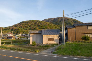 Ahşap kırsal evler, sıcak akşam güneşi altında yemyeşil tepeler tarafından desteklenen sessiz bir yol kenarında dururlar. Sahne, doğal dokular ve yumuşak gölgelerle huzurlu bir köy atmosferini yakalıyor..
