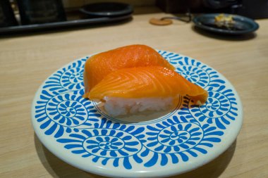 Bir çift somon nigiri suşi beyaz bir tabakta koyu mavi geometrik desenlerle sunuluyor. Çağdaş bir Osaka suşi restoranında hafif ahşap bir tezgahın üzerine kurulmuş..