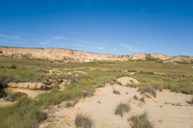 Düşük kum tepecikleri, engin yeşil bozkırlara ve Bardenas Reales, İspanya 'da canlı mavi bir gökyüzünün altında çarpıcı bej kayalıklara yol açar. Sahne açık, güneşli ve doğal toprak dokularıyla huzurlu..
