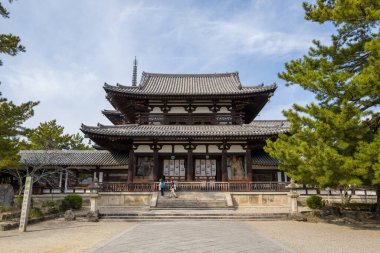 Japonya 'nın Nara kentindeki Horyuji tapınağındaki büyük Chumon Kapısı' nın önünde katmanlı kiremitli çatılar, ahşap sütunlar ve girişi çevreleyen muhafız heykelleri bulunuyor. Yeşil çam ağaçlarıyla çevrili tarihi yapı parlak bir gökyüzünün altında duruyor.