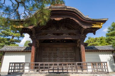 Kyoto 'daki Daitoku-ji Tapınağı' ndaki çam dallarının altında, parlak güneş ışığıyla aydınlanan kavisli çatılı ve karmaşık oymalarla süslü geleneksel ahşap bir kapı bulunur..