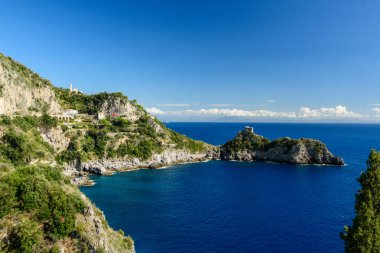 İtalya 'nın Capo di Conca kentindeki derin mavi suların üzerinde yükselen gür Akdeniz bitkileriyle kaplı kayalıklar. Tarihi bir gözetleme kulesi, canlı gökyüzü ve Amalfi Sahili boyunca ışıl ışıl deniz tarafından çerçevelenmiş küçük bir promoner üzerinde duruyor..