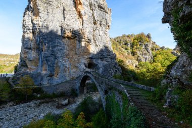 Tarihsel taş kemer köprüsü, Yunanistan 'ın Epirus kentindeki Pont Gefiri Kokkorou kenti yakınlarındaki yüksek kireçtaşı uçurumunun altındaki kurumuş bir nehir yatağı boyunca kıvrımlar. Parlak güneş ışığı, yapılandırılmış duvarcılık ve dramatik kanyon manzarasını ön plana çıkarıyor.