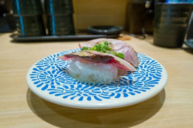 Çağdaş bir Osaka suşi restoranının soluk ahşap tezgahında sunulan, beyaz bir tabağın üzerinde yeşil soğanla süslenmiş bir parça nigiri suşi duruyor..