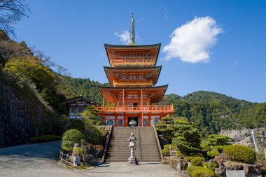Japonya 'nın Wakayama eyaletinin Kumano Nachi Taisha kentindeki canlı, üç katlı bir pagoda' nın önü. Heykel yeşillik tarafından çerçevelenmiş ve tek bir bulutla birlikte açık mavi bir gökyüzünün altındaki orman dağları. Güneş ışığı mimari ayrıntıları ve bereketli manzarayı vurgular.