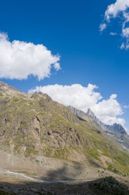 Dik, engebeli yamaçlar, yeşillik desenleri, parlak mavi gökyüzünün altında yükseliyor, büyük beyaz bulutlarla benekli. Güneş ışığı ve gölgeler, dağlık arazinin dramatik dokusunu vurgular..