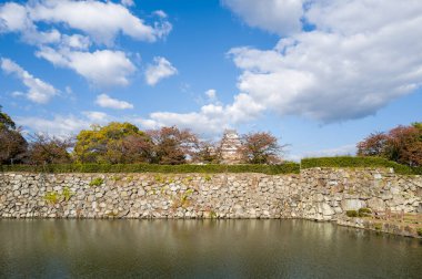 Tarihi taş duvarlar ve sakin bir hendek Japonya 'nın Himeji Kalesi' ndeki gökyüzünü yansıtır. Beyaz kale kulesi sonbahar ağaçlarının üzerinde kısmen görülebilir. Parlak gün ışığı ve dağınık bulutlar dingin ve zamansız bir atmosfer yaratır..