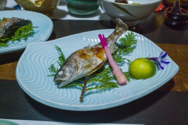 Taze yeşil yapraklı mavi dalga desenli bir tabakta ızgara nehir balığı servis edilir. Yuvarlak bir narenciye, pembe garnitür ve mor yenilebilir bir çiçek rafine edilmiş bir Japon sunumu yaratır..