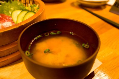 Dilimlenmiş soğanla süslenmiş bir kase geleneksel miso çorbası Japon restoranındaki sıcak ortam ışıklarıyla aydınlatılmış ahşap bir sashimi küvetinin yanında oturur..