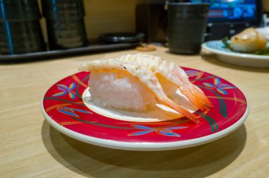 Çağdaş bir Osaka suşi restoranında açık ahşap tezgahta sergilenen canlı kırmızı bir tabakta kremalı mayonezle kaplı iki parça kurutulmuş karides nigiri sushi servis edilir..