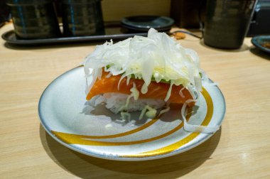 Avokado, ince dilimlenmiş soğanla kaplı tek bir parça somon balığı ve modern bir Osaka suşi restoranında gümüş ve altın desenli bir tabağın üzerine serpiştirilmiş bir mayonez. Sunumda taze dokular ve minimalist bir stil vurgulanıyor.