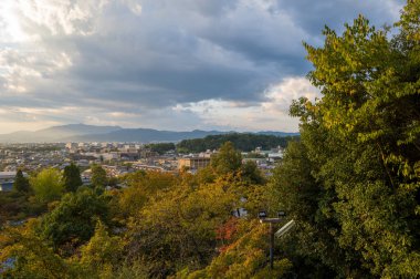 Renkli sonbahar yaprakları, yemyeşil ağaçlar ve dramatik bulutlar ve yumuşak altın ışıkla dolu gökyüzünün altındaki uzak dağlarla Kyoto şehrinin manzarası yükseldi..