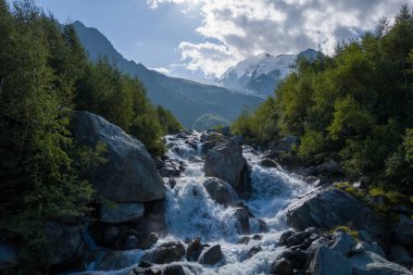 Alelacele akan dere, yemyeşil ağaçlar arasında kayalar üzerine yuvarlanır. Arka planda dramatik dağlar ve parçalı bulutlu bir gökyüzü vardır..