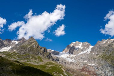 Buzul de la Lee Blanche ve engebeli Aiguilles de Tre la Tete 'nin çarpıcı manzarası. Kayalık yamaçlar, buzlu dokular ve kabarık mavi bulutlarla kaplı yeşil çimenler..