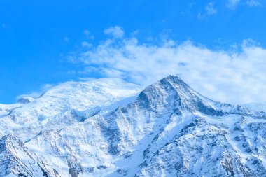 Karla kaplı Mont Blanc ve Aiguille du Gouter parlak mavi bir gökyüzü altında heybetli bir şekilde yükselirler. Kıtır kıtır dokular ve yumuşak bulutlar Alp manzarasına derinlik katar..