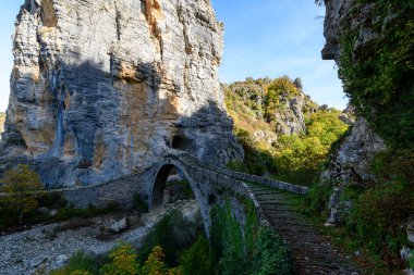 Historic stone arch bridge spans a rocky riverbed beneath dramatic limestone cliffs and lush autumn foliage near Pont Gefiri Kokkorou in Epirus, Greece. Soft daylight reveals textured masonry and the rugged natural landscape of the Vikos Gorge region