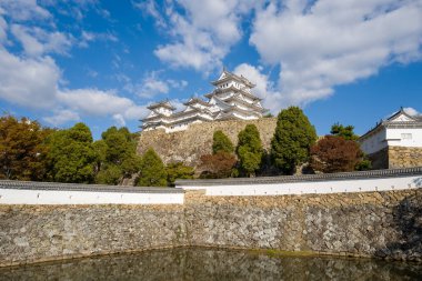 Himeji Kalesi 'nin zarif beyaz kuleleri devasa taş duvarların üzerinde yükseliyor ve gür yeşil ve sonbahar ağaçlarıyla süslenmiş, yumuşak bulutlarla benekli, sakin bir hendek..