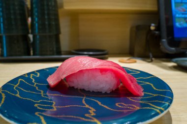 Osaka 'da modern bir suşi restoranında, açık ahşap bir tezgahın üzerine kurulmuş, parlak mavi bir tabağa yerleştirilmiş tek bir parça ton balığı nigiri..