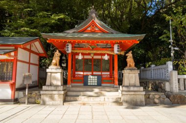 Japonya 'nın Kobe kentinde bulunan kompakt bir Shinto türbesi pavyonunda canlı kırmızı kirişler, yeşil kiremitli bir çatı ve girişte iki taş koruyucu köpek heykeli bulunur. Güneş ışığı mimari detayları vurgular ve asfalt avluda yumuşak gölgeler bırakır, yemyeşil