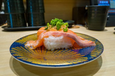 Kesilmiş yeşil soğanla kaplı bir parça somon nigiri modern bir Osaka suşi restoranında soluk bir ahşap tezgahta sergilenen dekoratif mavi ve altın bir tabakta duruyor..