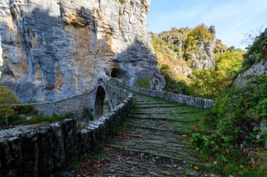 Antik taş kemer köprüsü, Yunanistan 'ın Epirus kentindeki Vikos Vadisi bölgesinde dramatik kireçtaşı uçurumlarının altında kayalık bir nehir yatağını ve gür sonbahar yapraklarını geçer. Yumuşak gün ışığı desenli duvarcılığı, canlı yeşilliği ve engebeli doğal manzarayı gözler önüne seriyor..
