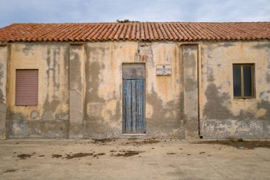 Sardalya 'daki Asinara Ulusal Parkı' nda soyulmuş sarı alçı, soluk mavi ahşap kapı ve terasotta çatı kiremitleriyle yıpranmış bir cephe duruyor. Sessiz dokular ve bulutlu gökyüzü ihmalkarlık hissi uyandırıyor..