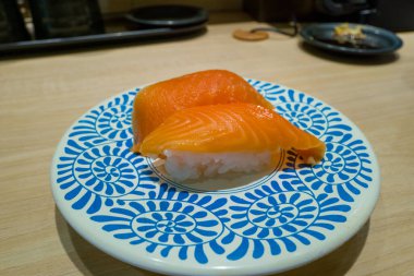 İki parça parlak somon nigiri suşi koyu mavi spiral desenli beyaz bir tabakta duruyor. Çağdaş bir Osaka suşi restoranının soluk ahşap tezgahında sergileniyor..