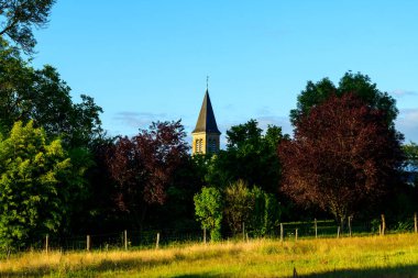 Sivri uçlu bir kilise kulesi canlı yeşil ve bordo ağaçların arkasında yükseliyor. Güneşli çimenli bir tarlası ve Saint-Honore-les-Bains 'de açık mavi gökyüzü var..
