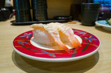 Kremalı mayonezle kaplanmış iki parça kurutulmuş karides nigiri suşisi, çağdaş bir Osaka suşi restoranında açık ahşap bir tezgahın üzerine kurulmuş mavi ve altın çiçekli bir tabakta sunuluyor..