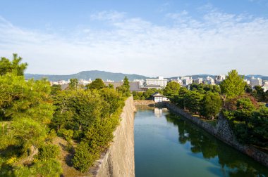 Tarihi taş duvarlar ve yemyeşil yeşilliklerle kaplı huzurlu bir hendeğin yüksek perspektifi, Kyoto şehri ve parlak mavi gökyüzünün altındaki uzak dağlar.