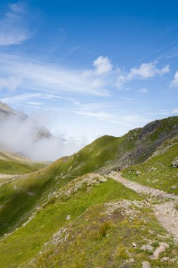 Beaufortain Alpleri 'ndeki yemyeşil yamaçlar boyunca kayalık bir yürüyüş yolu kıvrılır. Sürüklenen bulutlar ve canlı mavi bir gökyüzü taze, açık bir dağ atmosferi yaratır..