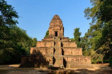 Phnom Bakheng 'in antik kumtaşı piramidi, Angkor, Kamboçya' da yemyeşil tropikal ağaçlar ve açık mavi bir gökyüzü tarafından çerçevelenen orman zemininden yükselir. Kıtır güneş ışığı, bu tarihi Khmer anıtının yıpranmış duvarcılığını ve geometrik çizgilerini vurguluyor..